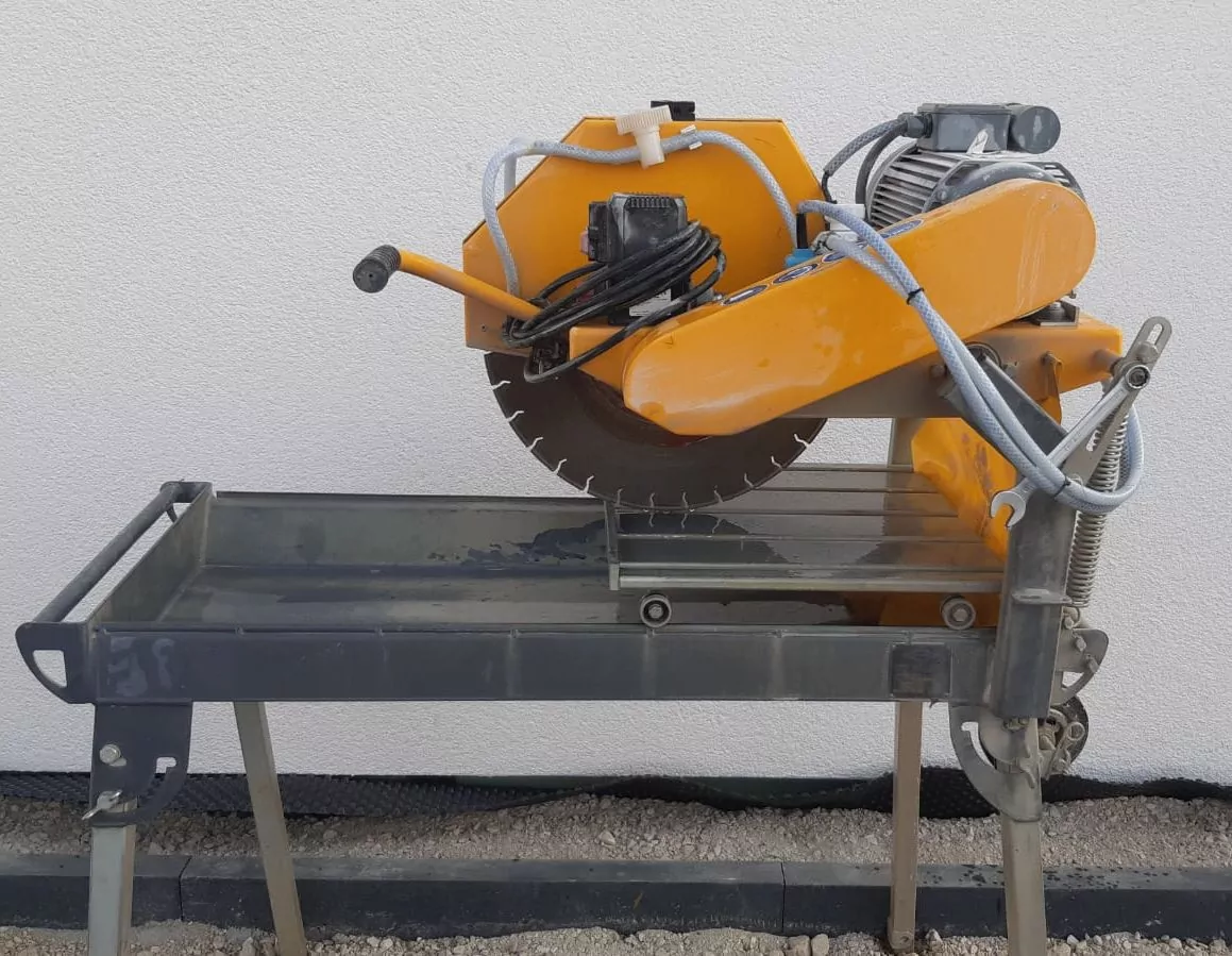 Eine große orangefarbene Steinsäge steht auf einem Metallständer vor einer weißen Wand. Die Säge hat ein rundes Sägeblatt, von dem Stromkabel ausgehen. Der Ständer steht auf einer Beton- und Kiesfläche.