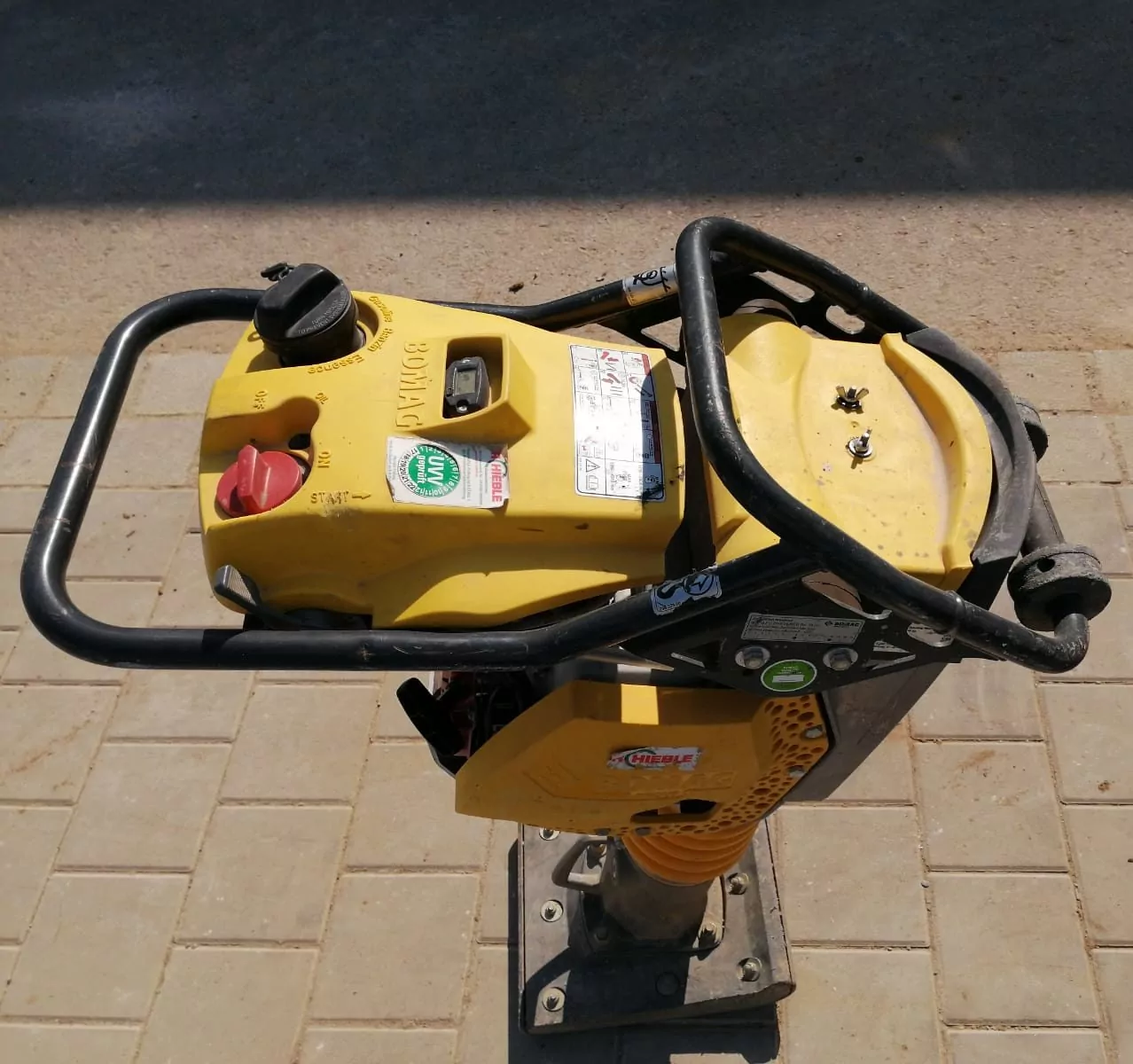 Eine Draufsicht auf eine gelbe Verdichtungsmaschine auf einer gepflasterten Fläche. Die Maschine hat einen runden Tankdeckel, Bedienhebel und verschiedene Etiketten und Aufkleber. Der Boden darunter besteht aus hellbraunen rechteckigen Ziegeln.