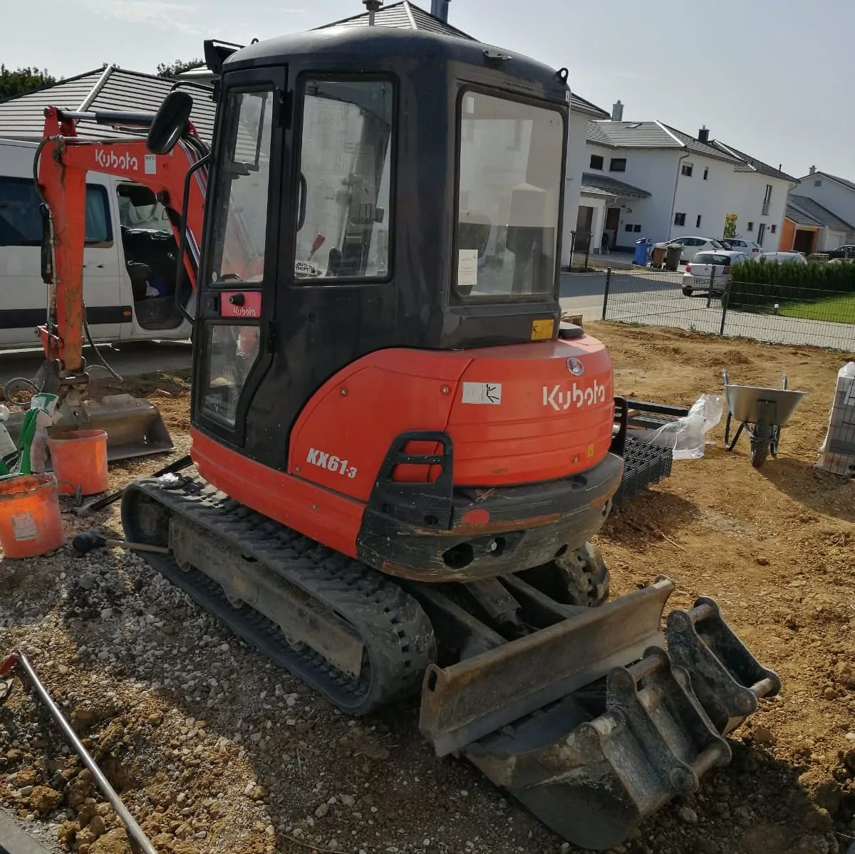 Ein kompakter rot-schwarzer Bagger Kubota KX61-3 steht auf einer Baustelle, die Schaufel ruht auf dem Boden. Er ist umgeben von Erde, Baumaterialien und im Hintergrund sind Wohnhäuser zu sehen.