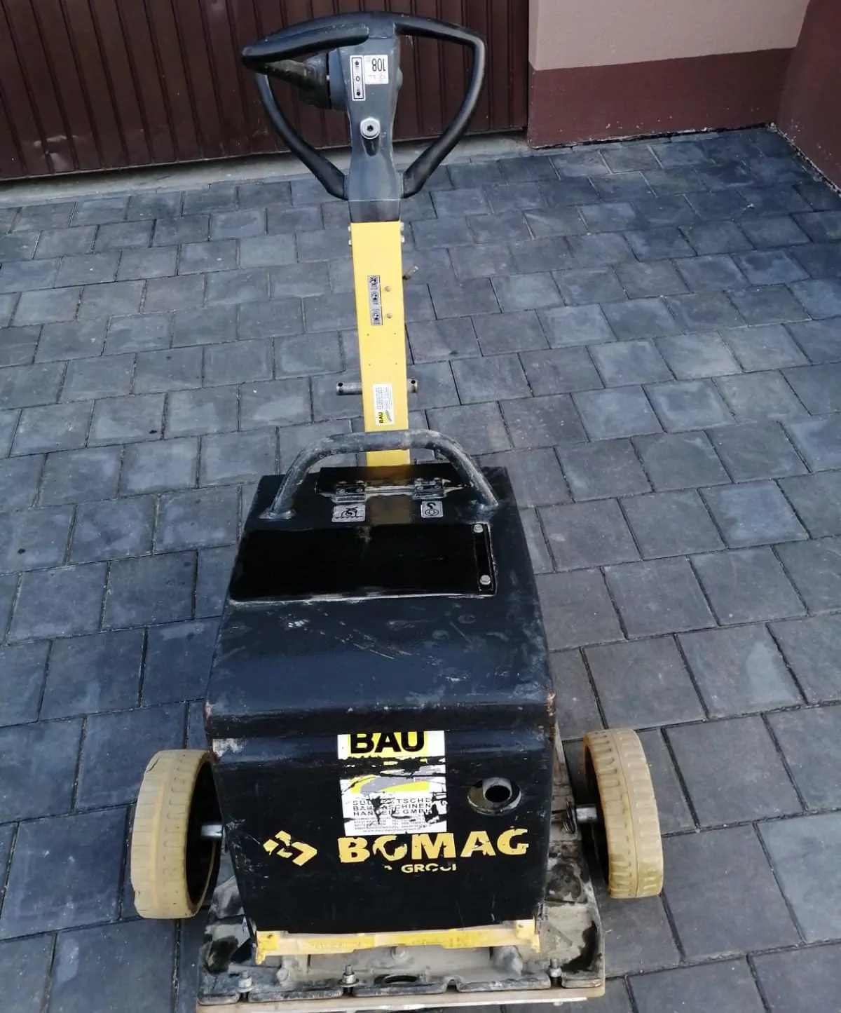 Eine gelb-schwarze Bomag-Rüttelplatte steht auf einem grauen Ziegelpflaster. Der Griff steht aufrecht und die Maschine weist Gebrauchsspuren auf, auf der Oberfläche ist Schmutz sichtbar.