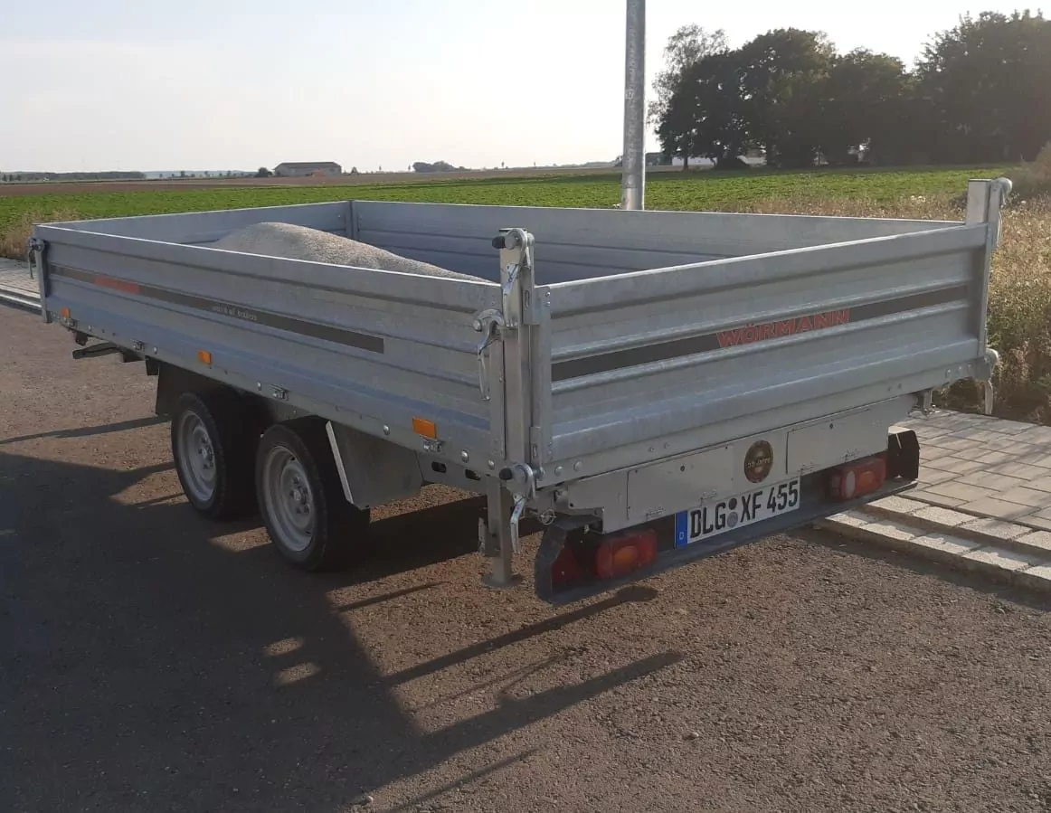 Ein kleiner Pritschenanhänger mit herunterklappbaren Seitenwänden steht auf einer asphaltierten Straße neben einem Feld. Der Anhänger ist teilweise mit Sand oder Kies beladen. Die Szene spielt bei Tageslicht und wirft Schatten auf den Boden.