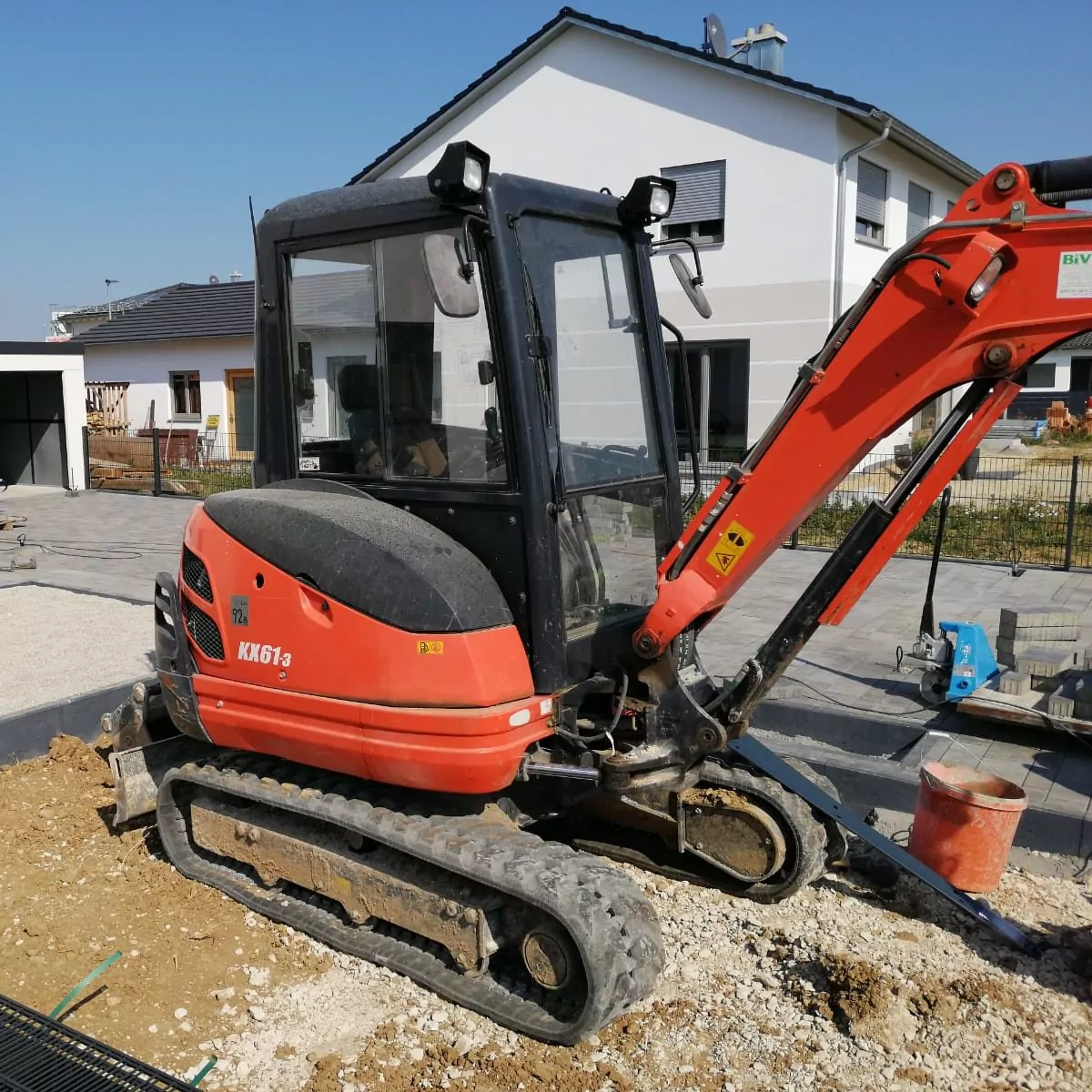 Ein kleiner rot-schwarzer Bagger steht auf einer Baustelle in der Nähe eines modernen weißen Hauses mit dunklem Dach. Die Maschine hat Gummiketten und steht auf unebenem Boden, in der Nähe liegen verstreute Baumaterialien.