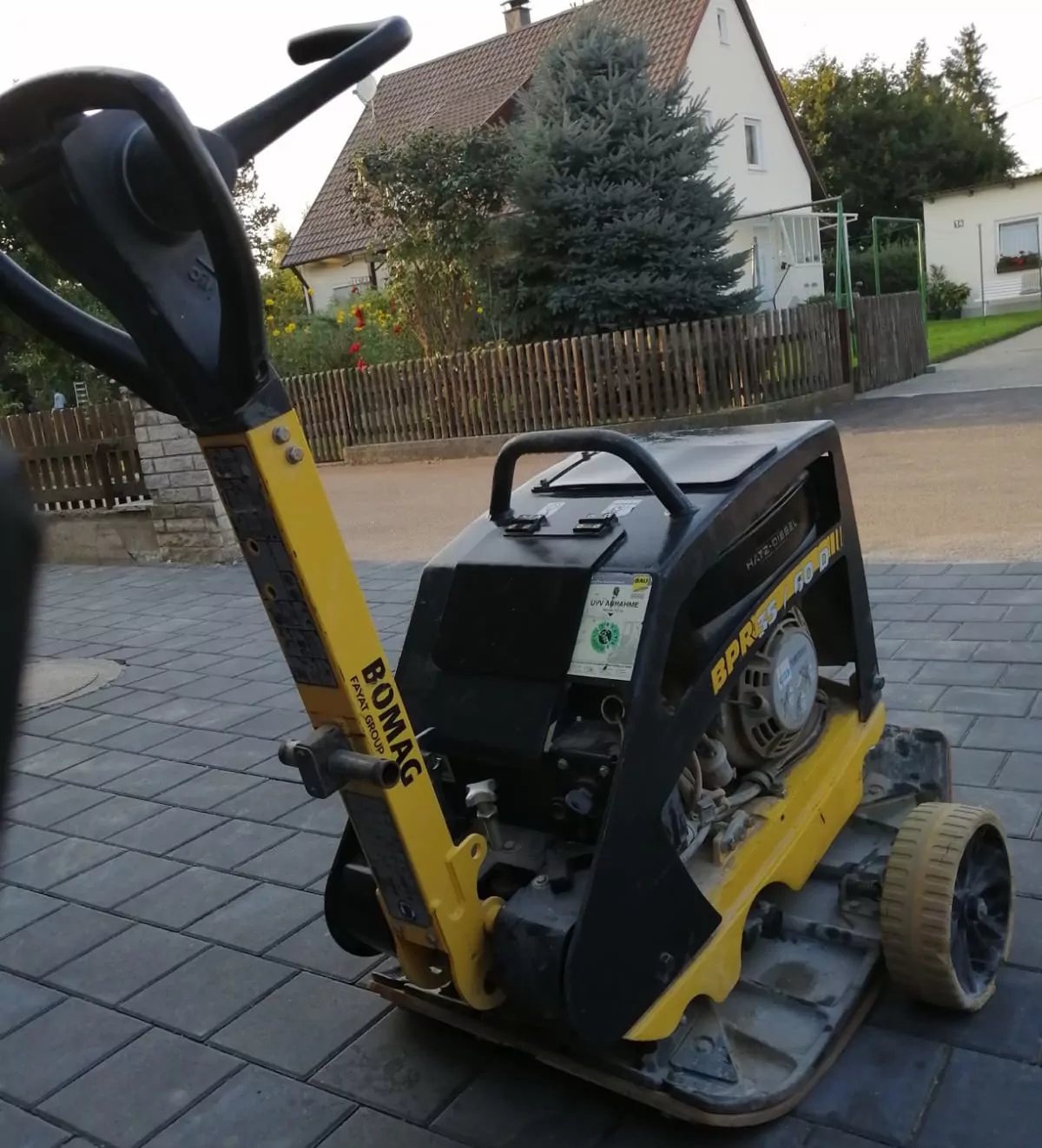 Eine gelb-schwarze Bomag-Rüttelplatte steht auf einer gepflasterten Einfahrt. Im Hintergrund ist eine Vorstadtgegend mit einem Haus, Bäumen und einem Garten zu sehen. Die Platte trägt ein Etikett mit der Modellnummer BPR 35/60 D.