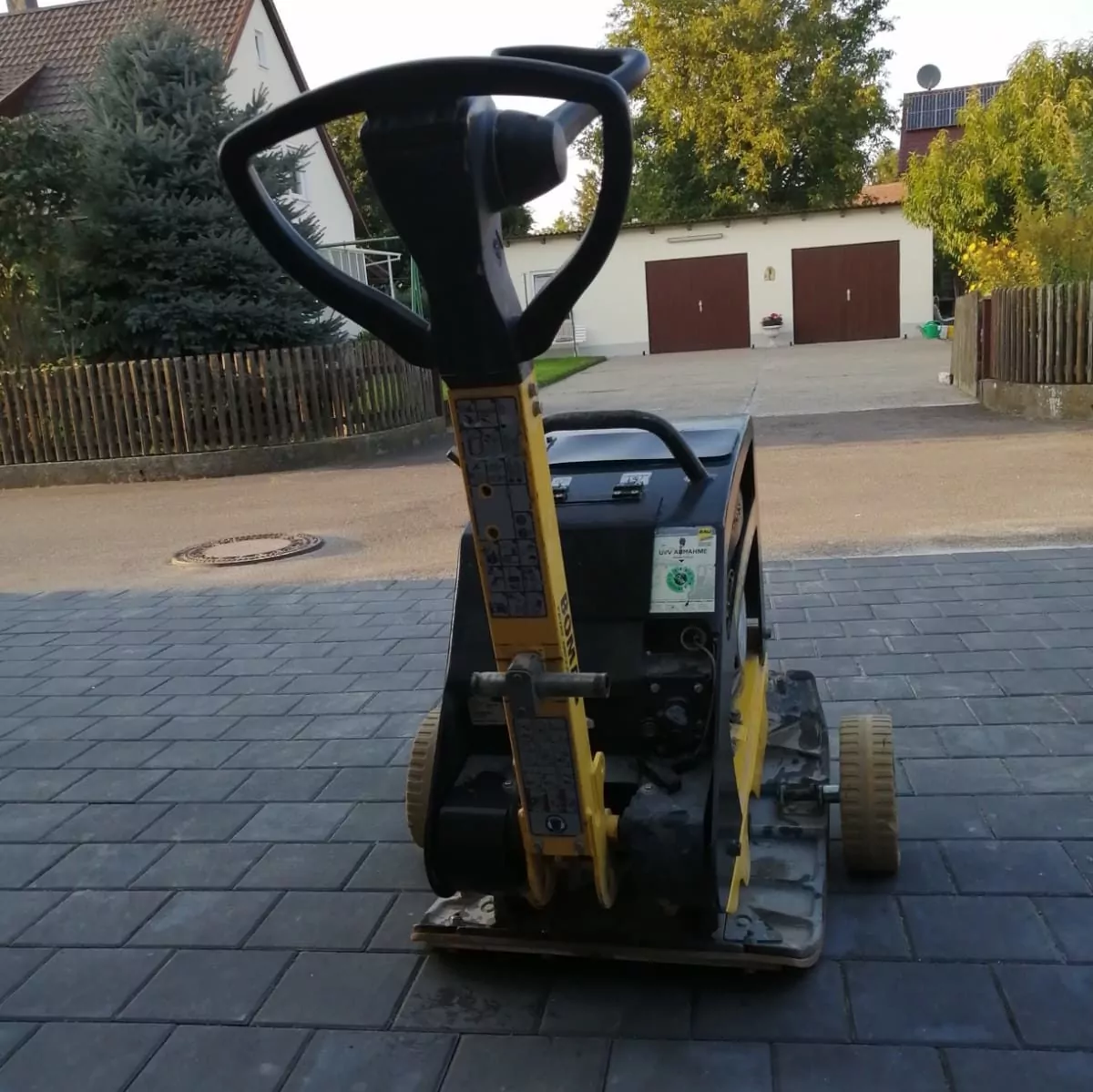 Eine Nahaufnahme eines kompakten Bodenverdichters auf einer gepflasterten Einfahrt. Die Maschine verfügt über einen Griff zur Bedienung und weist ein robustes Design auf, das für Bau- oder Landschaftsbauarbeiten geeignet ist. Im Hintergrund sind Wohnhäuser und Bäume zu sehen.