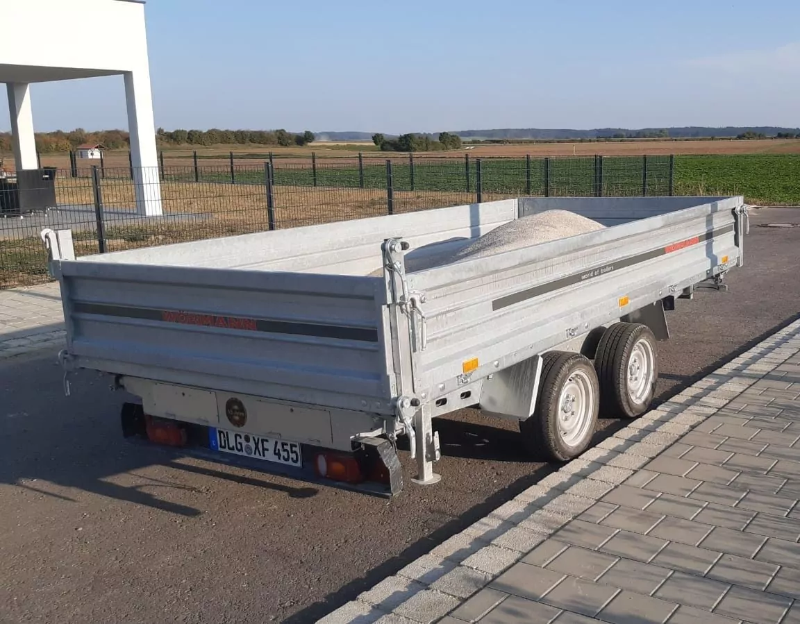 Ein grauer Anhänger mit Doppelachse steht auf einer befestigten Fläche. Der Anhänger ist teilweise mit Sand oder Kies gefüllt. Auf der Rückseite ist ein Nummernschild angebracht und im Hintergrund ist ein Feld mit Bäumen zu sehen.
