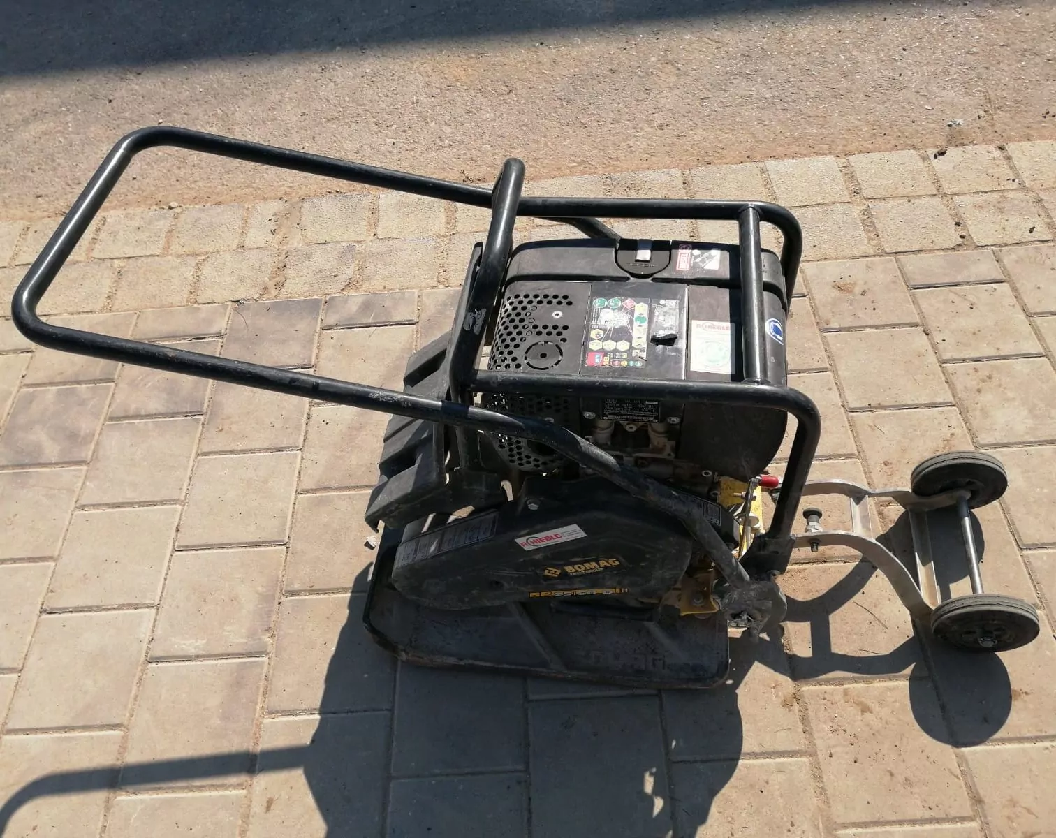 Eine schwarze Rüttelplatte mit stabilem Metallrahmen steht auf einer befestigten Fläche. Die Maschine verfügt über Handgriffe zum Manövrieren und sichtbare Bedienelemente auf der Oberseite. Ihr Schatten wird durch das Sonnenlicht auf die Straße geworfen.