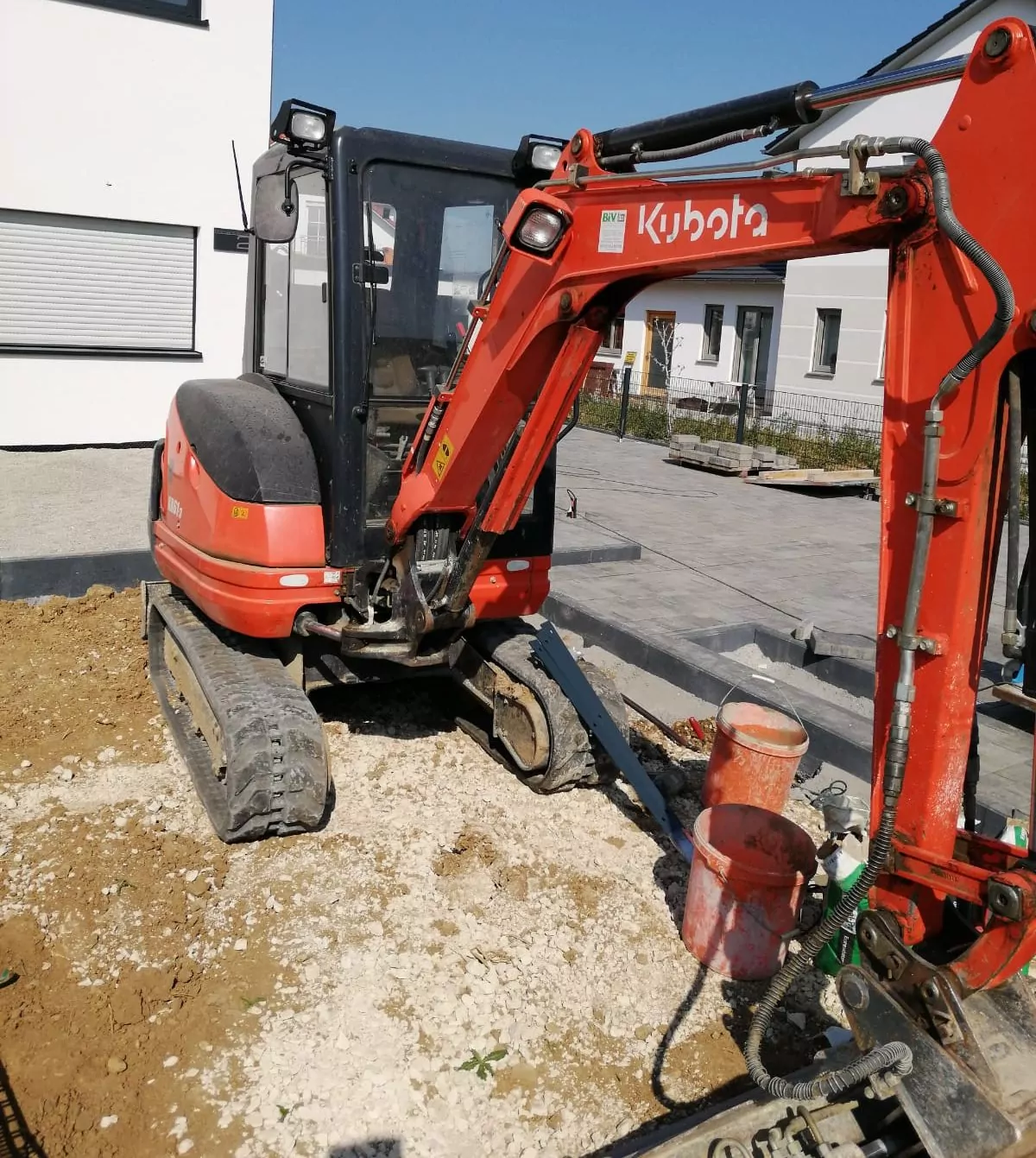 Ein roter Kubota-Minibagger steht auf einer Baustelle. Er hat eine Frontschaufel und ist von Erde und Kies umgeben. In der Nähe stehen eine rote Schaufel und andere Baumaterialien. Im Hintergrund sind zwei weiße Gebäude und ein klarer blauer Himmel zu sehen.
