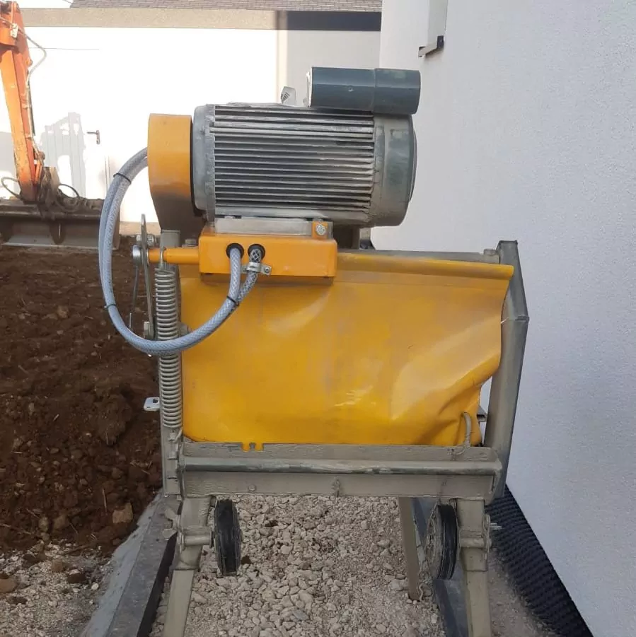 Ein großer Industriemotor ist auf einem gelben Metallgehäuse montiert, an dem ein flexibler Schlauch befestigt ist. Die Einheit befindet sich auf einer Baustelle mit einer Kiesfläche und einem unfertigen Gebäude im Hintergrund.