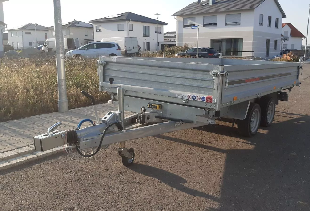 Ein Metallanhänger mit zwei Achsen steht auf einer befestigten Fläche in einem Wohngebiet. Er hat eine Anhängerkupplung, eine Kurbel und eine Seilwinde an der Vorderseite. Im Hintergrund sind mehrere Häuser und geparkte Autos zu sehen.