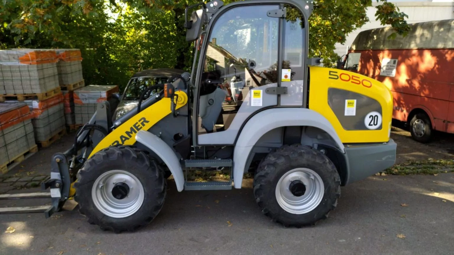Ein gelb-grauer Kramer 5050-Lader steht auf einer gepflasterten Fläche. Er hat vier große Reifen und eine geschlossene Kabine. Im Hintergrund sind Stapel von Baumaterialien und ein roter Lieferwagen neben Bäumen zu sehen.