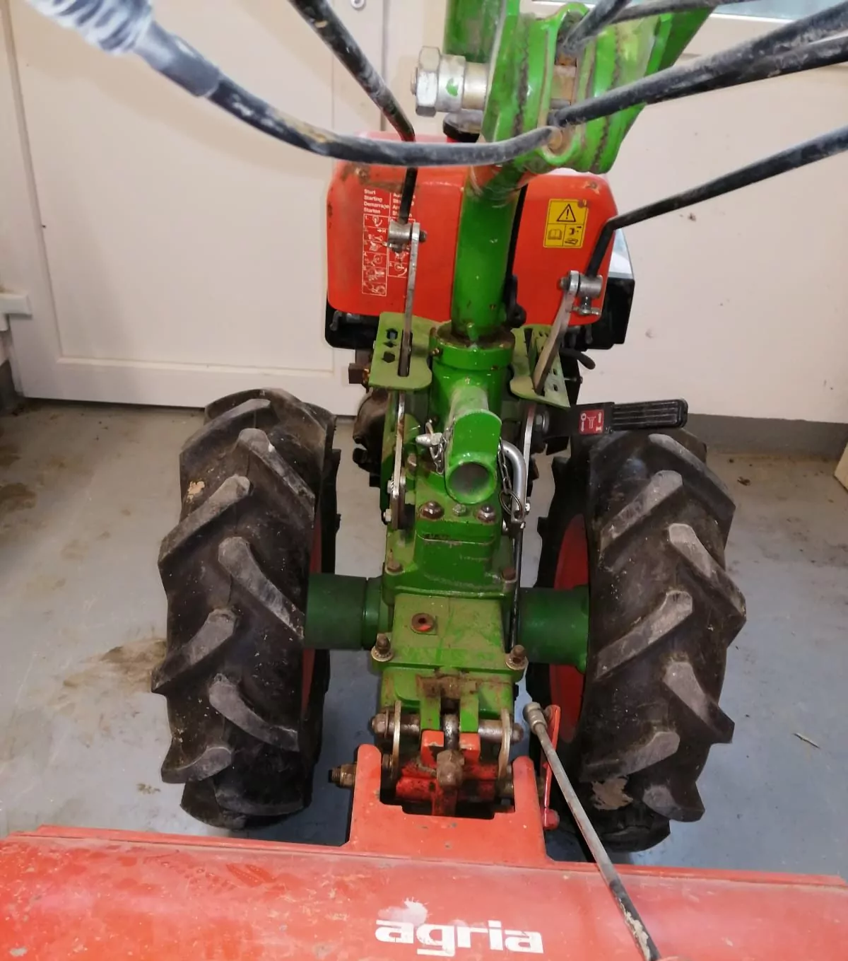 Eine rot-grüne Fräse mit großen schwarzen Reifen steht drinnen auf einem grauen Boden. Das Gerät verfügt über einen Lenker mit schwarzen Kabeln und einen vorderen Metallaufsatz. Auf dem Aufsatz ist der Markenname „agria“ zu sehen.