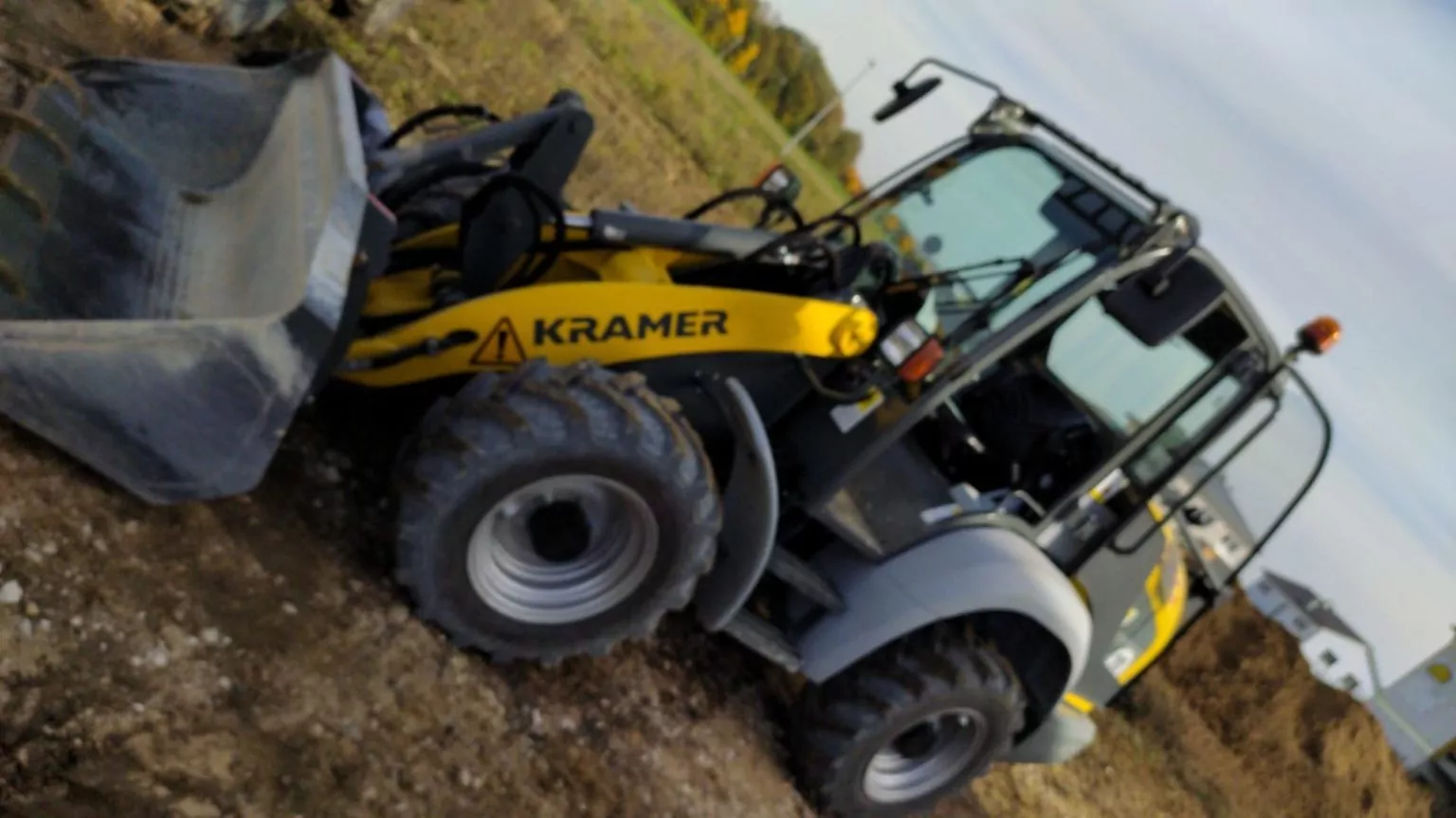 Ein gelb-grauer Kramer-Frontlader steht auf einem unbefestigten Gelände. Das Fahrzeug hat große Räder und eine Frontschaufel. Im Hintergrund sind Grasflächen und ein klarer Himmel zu sehen.
