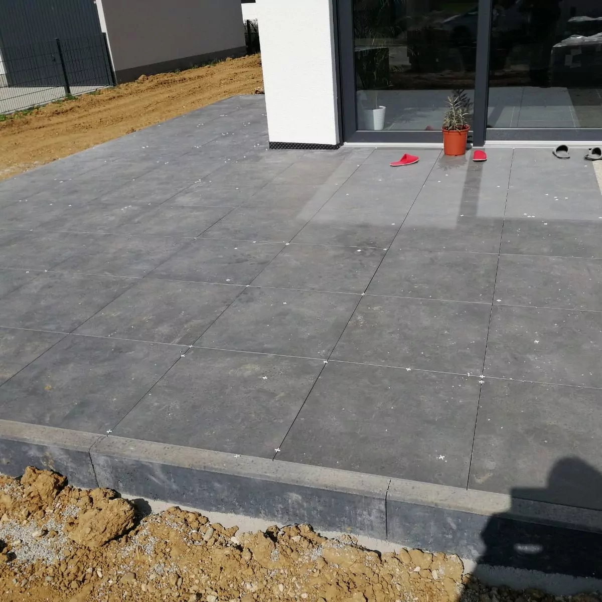 Von einem Gebäude aus erstreckt sich eine Terrasse mit großen grauen Fliesen. Ein Paar rote Sandalen und eine Pflanze in einem Topf stehen neben den Glasschiebetüren. Der Boden um die Terrasse herum ist kahl und im Vordergrund ist ein Schatten zu sehen.