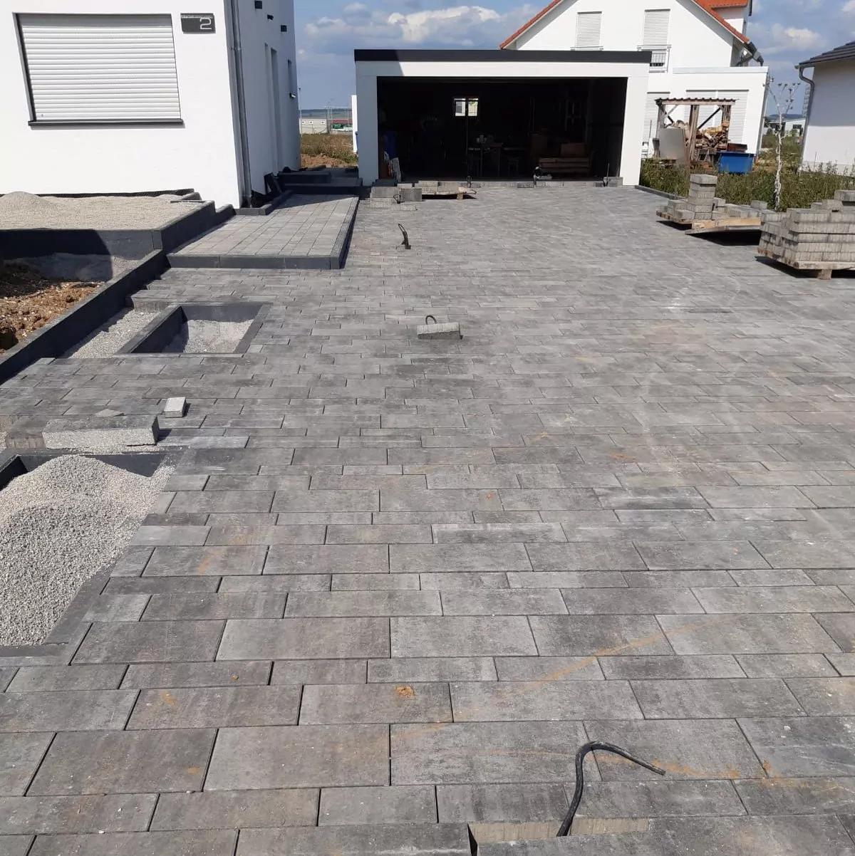 Eine im Bau befindliche Auffahrt mit grauen Pflastersteinen. Im Vordergrund sind teilweise verlegte Steine zu sehen, in einigen Bereichen mit losem Kies. Im Hintergrund sind eine Garage und ein weißes Gebäude zu sehen. Auf der rechten Seite sind Stapel mit Baumaterialien zu sehen.