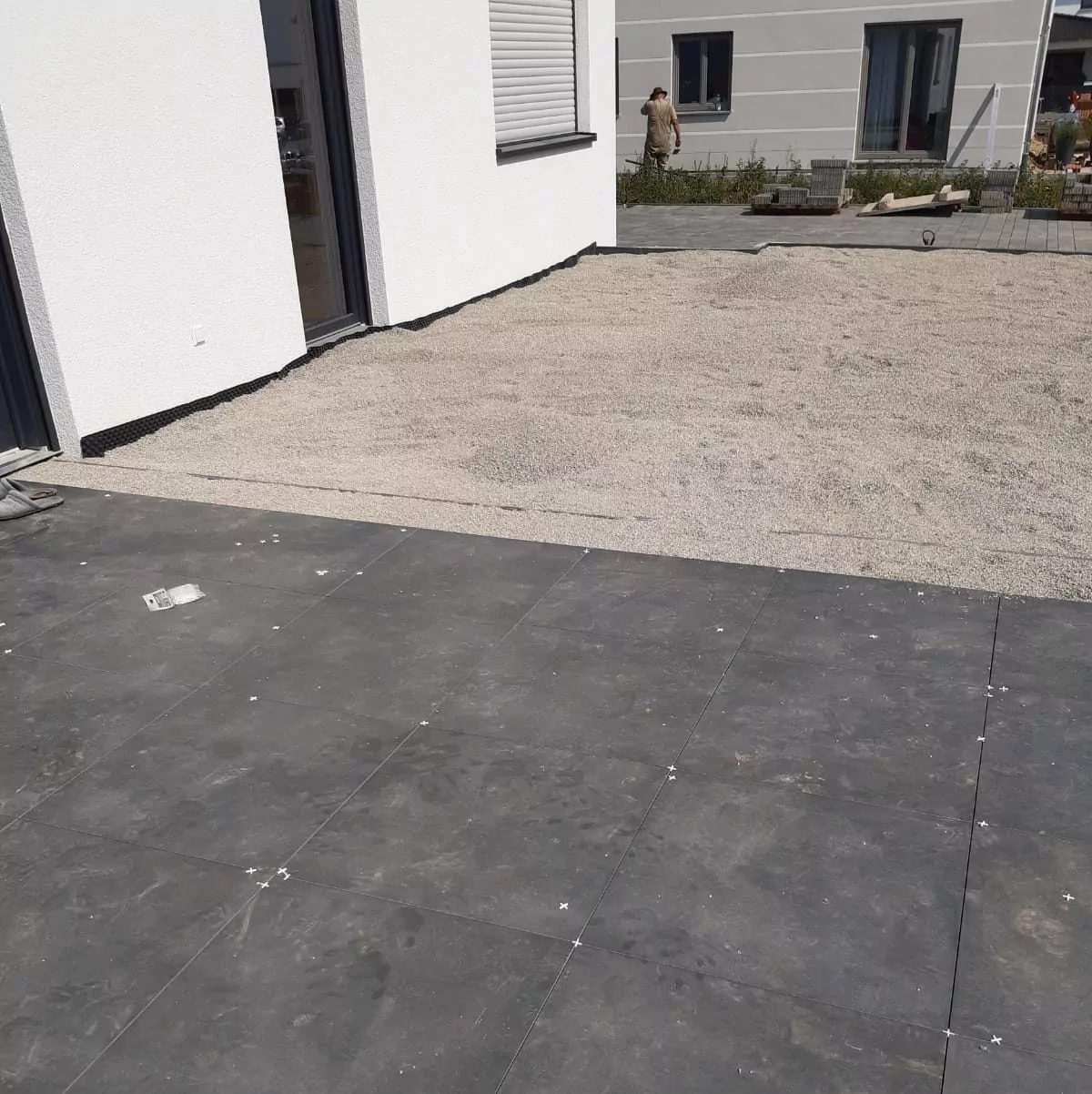 Ein Terrassenbereich wird gerade umgebaut. Im Vordergrund sind frisch verlegte dunkle Steinfliesen zu sehen, während im Hintergrund ein Abschnitt mit Kies zu sehen ist. Eine teilweise sichtbare Person arbeitet in der Nähe eines modernen Gebäudes. Werkzeuge und Materialien liegen verstreut herum.
