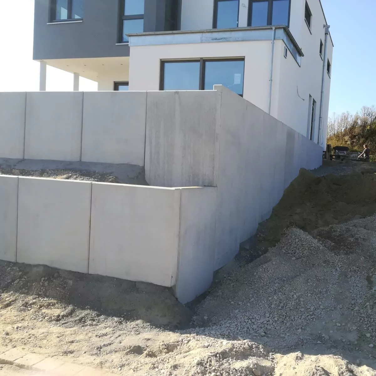 Ein modernes weißes Gebäude mit großen Fenstern steht neben einer Stützmauer aus grauem Beton. Die Mauer steht an einem Erdhang und auf dem Boden sind einige Baumaterialien sichtbar. Der Himmel ist klar und die Szene wird vom Sonnenlicht erhellt.