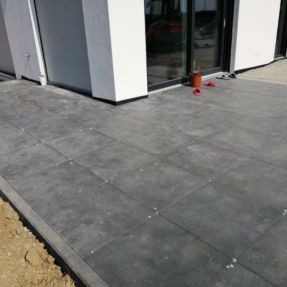Gezeigt wird ein modernes Gebäude mit großen Fenstern. Der Bereich verfügt über eine Außenterrasse mit grauen quadratischen Fliesen. In der Nähe des Eingangs stehen ein kleiner roter Blumentopf und rote Schuhe. Der Boden neben den Fliesen ist nackte Erde.