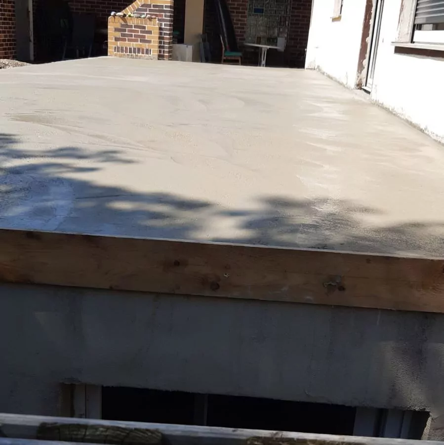Frisch gegossene Betonplatte mit glatter Oberfläche, eingefasst von einem Holzrahmen. Schatten von nahe gelegenen Objekten fallen auf den Beton. Im Hintergrund ist ein weißes Backsteingebäude zu sehen, darunter ein teilweise geöffnetes Fenster.