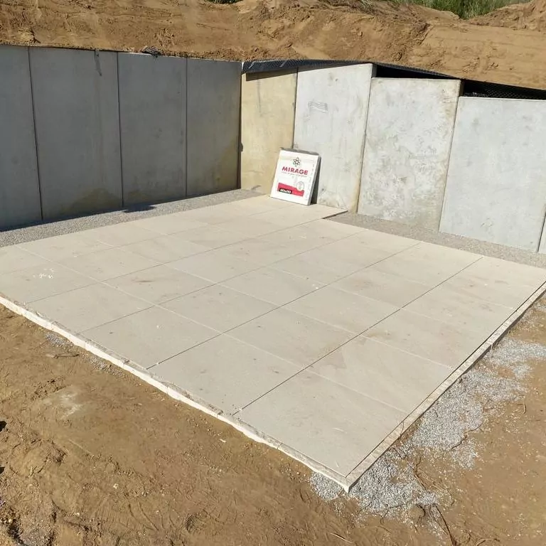 Eine Baustelle mit einem Abschnitt aus sauber verlegten weißen Fliesen auf dem Boden, eingefasst von Betonplatten und umgeben von Erde. Ein Paket mit der Aufschrift „Mirage“ lehnt an einem Erdwall.