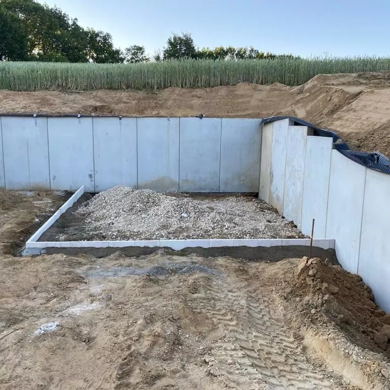 Eine Baustelle mit offenem Fundament, Betonwänden und Kiesboden. In der Umgebung ist der Boden freigelegt und man sieht Reifenspuren. Im Hintergrund sind eine Wiese und eine Reihe Bäume unter einem klaren blauen Himmel zu sehen.