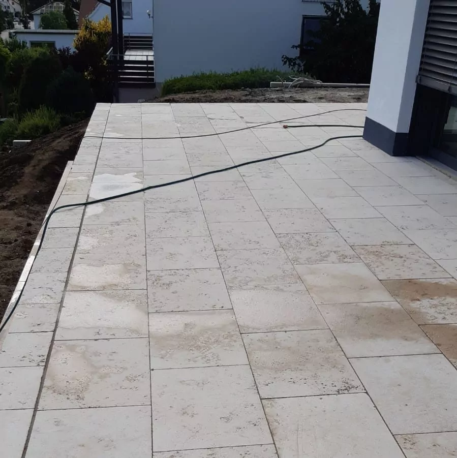 Großzügiger Außenterrassenbereich mit hellen, rechteckigen Fliesen. Ein grüner Gartenschlauch liegt quer über den Fliesen. Um die Terrasse herum ist ein Streifen Erde, im Hintergrund ist etwas Grün zu sehen. Ein modernes weißes Gebäude grenzt an.