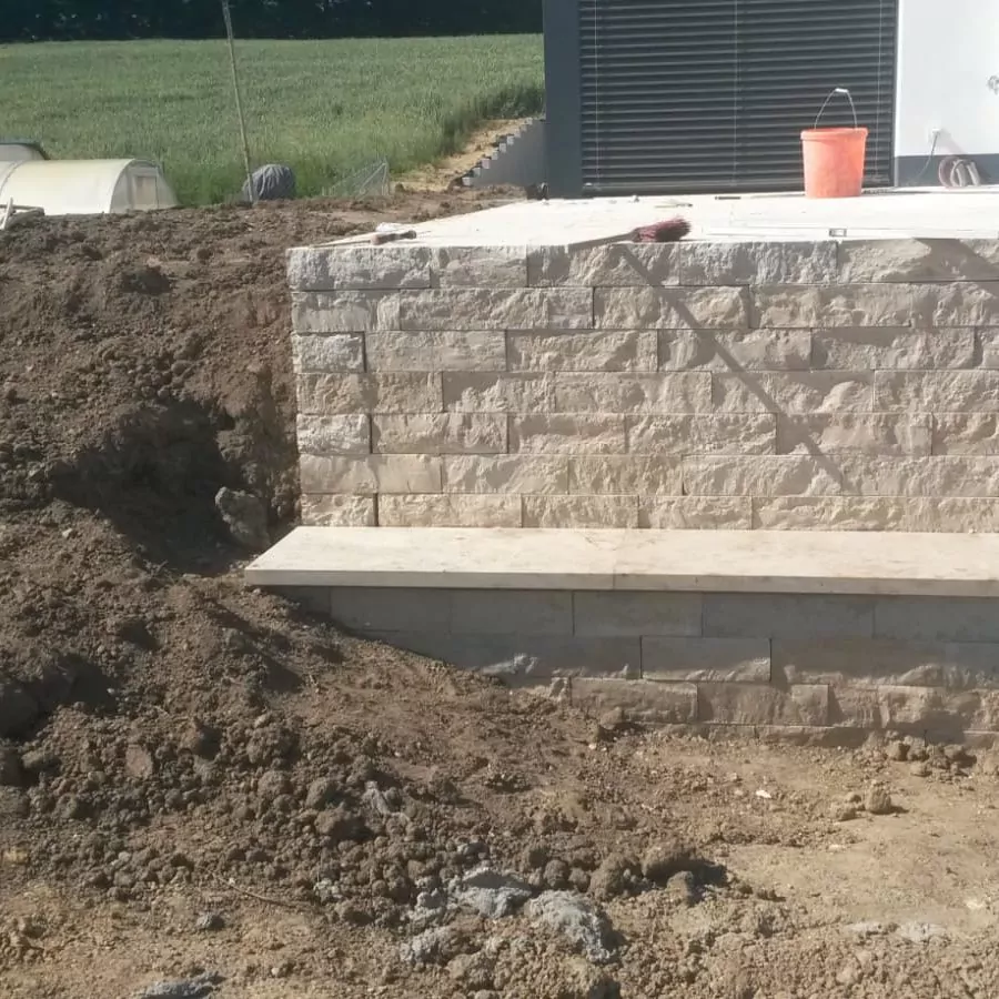 Bauszene mit einer Steinmauer, vor der Erde aufgehäuft ist. Von der Mauer geht eine flache Fläche ab, möglicherweise ein Sitzbereich. Auf der Mauer stehen ein roter Eimer und ein Werkzeug. Im Hintergrund sind ein Feld und ein Teil eines Gebäudes zu sehen.