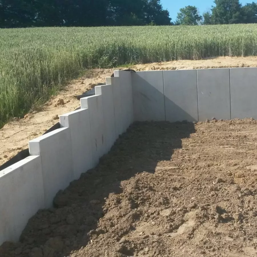 Betonstützmauer im Bau mit Erdauffüllung. Stufen an der Mauer führen hinauf zu einer Wiese. Im Hintergrund sind klarer blauer Himmel und Bäume zu sehen.