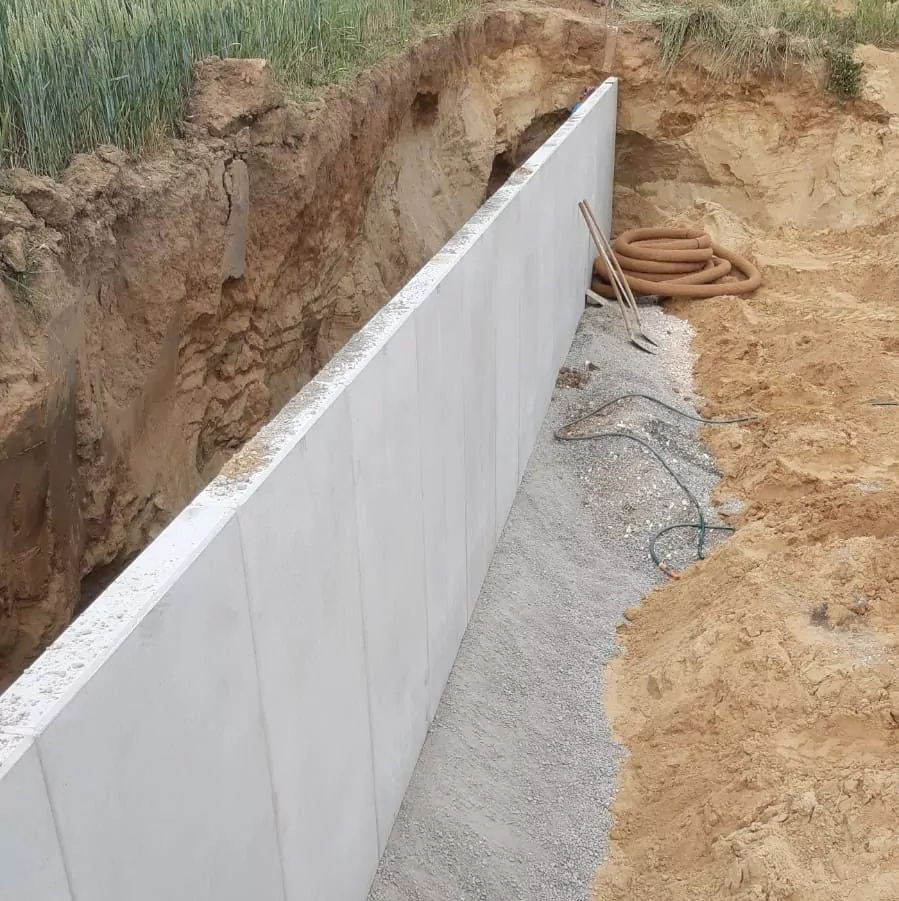 Eine neu errichtete Stützmauer aus Beton befindet sich in einem Graben, dessen eine Seite freiliegende Erde aufweist. In der Nähe liegen aufgerollte Schläuche, loser Kies und Werkzeuge verstreut auf der Baustelle.
