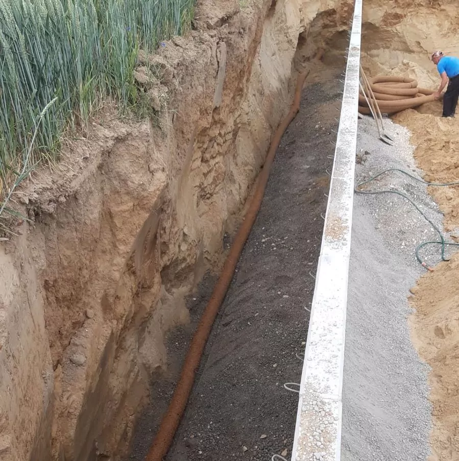Gezeigt wird ein tiefer Graben mit einem Drainagerohr am Boden. Der Graben ist teilweise mit Kies gefüllt. Eine Person steht am Rand und arbeitet mit Werkzeugen. Auf der linken Seite des Bildes ist ein Feld mit Nutzpflanzen zu sehen.