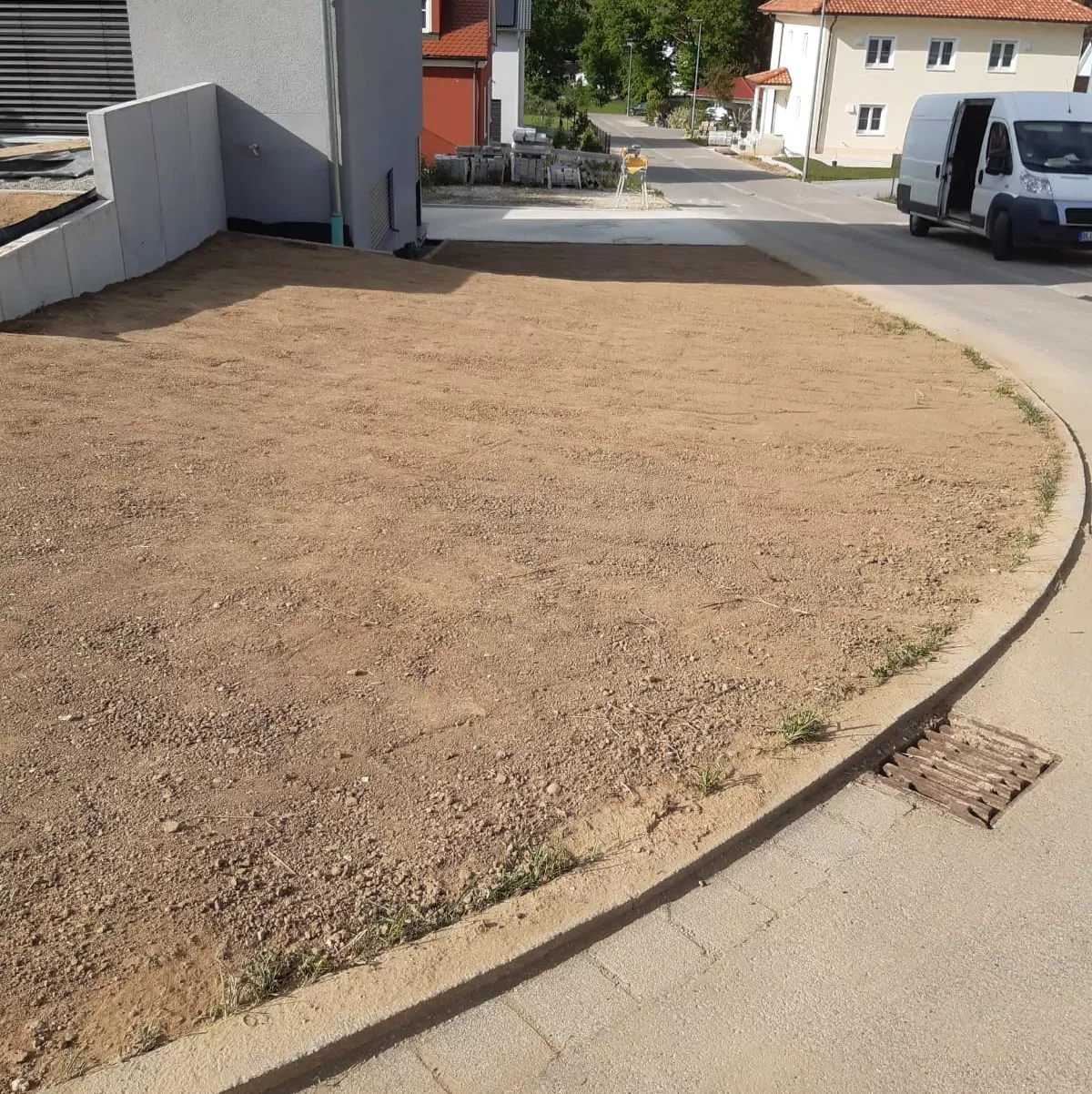 An einer Straßenecke eines Wohnhauses liegt ein mit Erde bedeckter Vorgarten neben einer asphaltierten Straße. Im Hintergrund sind mehrere Häuser zu sehen. Auf der rechten Straßenseite parkt ein weißer Lieferwagen. In der Nähe des Bordsteins befindet sich ein Abwasserkanal.