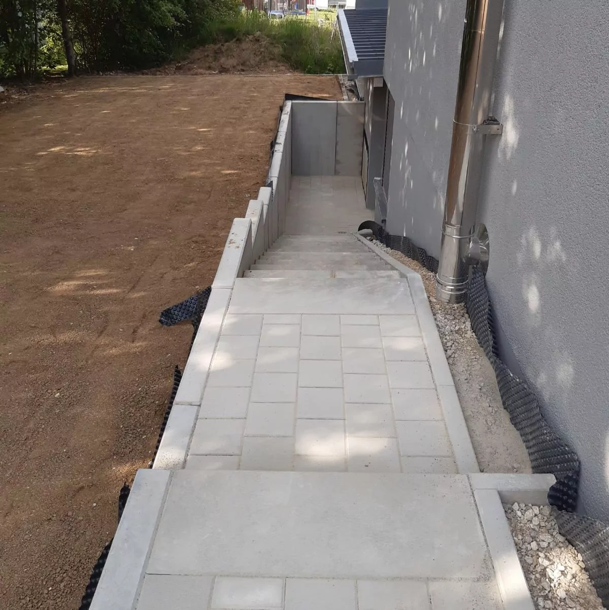 Eine neu errichtete Außentreppe mit hellen Fliesen verläuft neben einem Gebäude mit grauer Wand. Die Treppe ist durch Betonkanten begrenzt und führt auf eine niedrigere Ebene. Der umliegende Bereich ist mit Erde und Vegetation bedeckt.