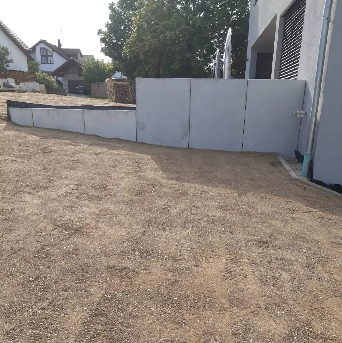Eine Kiesauffahrt führt zu einem hellgrauen Gebäude mit modernem Design. An der Seite des Gebäudes verläuft eine Betonmauer. Im Hintergrund sind ein Holzzaun und einige Bäume sowie ein Nachbarhaus zu sehen.