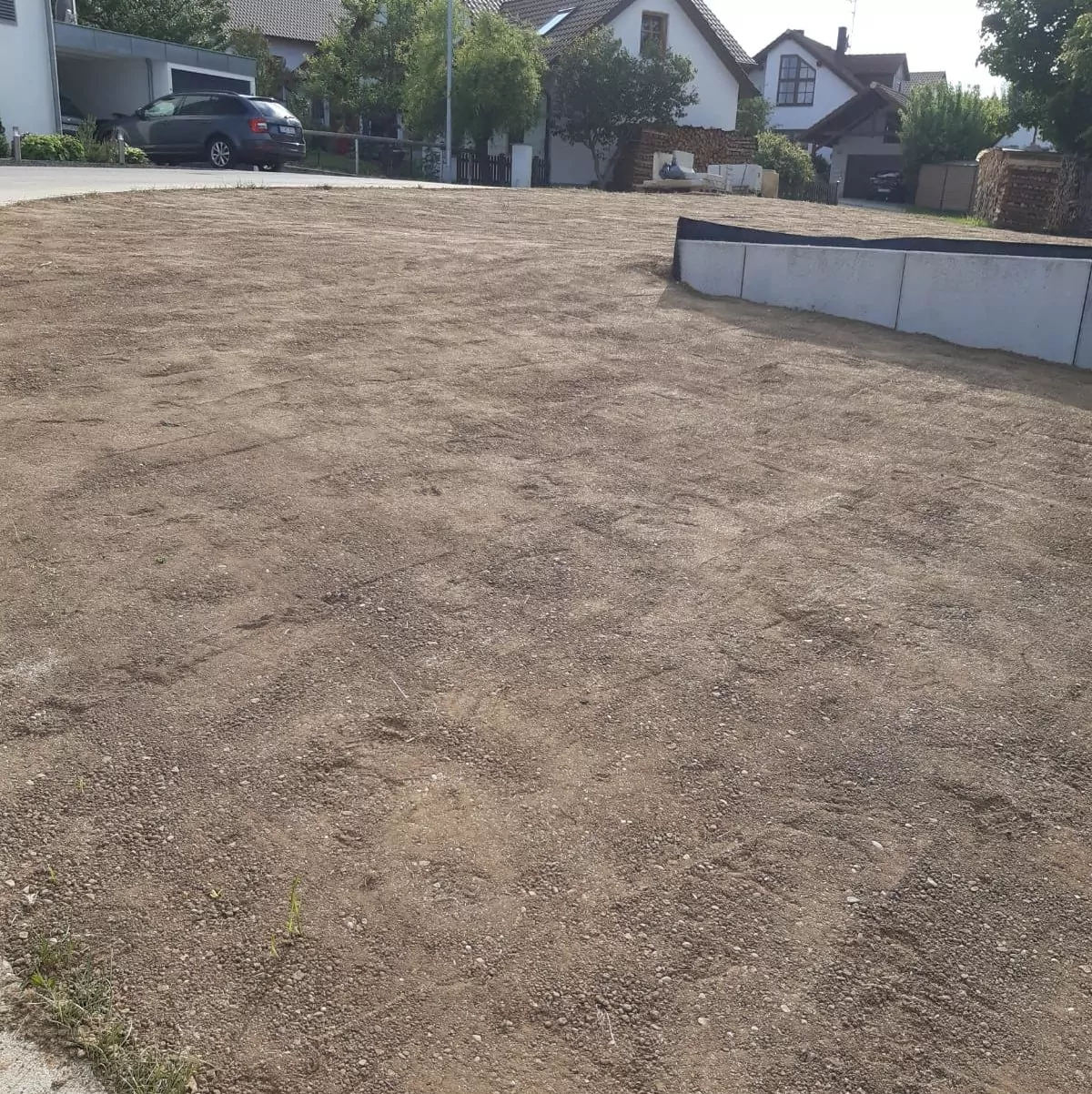 Ein unbefestigtes Grundstück in einem Wohngebiet mit ein paar Häusern und einem geparkten Auto im Hintergrund. Der Boden ist größtenteils kahl, mit einer kleinen erhöhten Betonstruktur auf der rechten Seite. In der Nähe der Gebäude sind Bäume und Sträucher sichtbar.