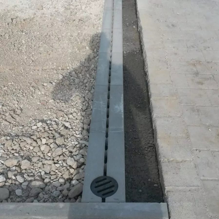 Ein Beton-Abflusskanal mit Schlitzabdeckung verläuft rechts neben einer gepflasterten Fläche. Auf der linken Seite sind ein Kieshaufen und blanker Boden zu sehen. Im Vordergrund ist ein kreisrunder Abfluss zu sehen.