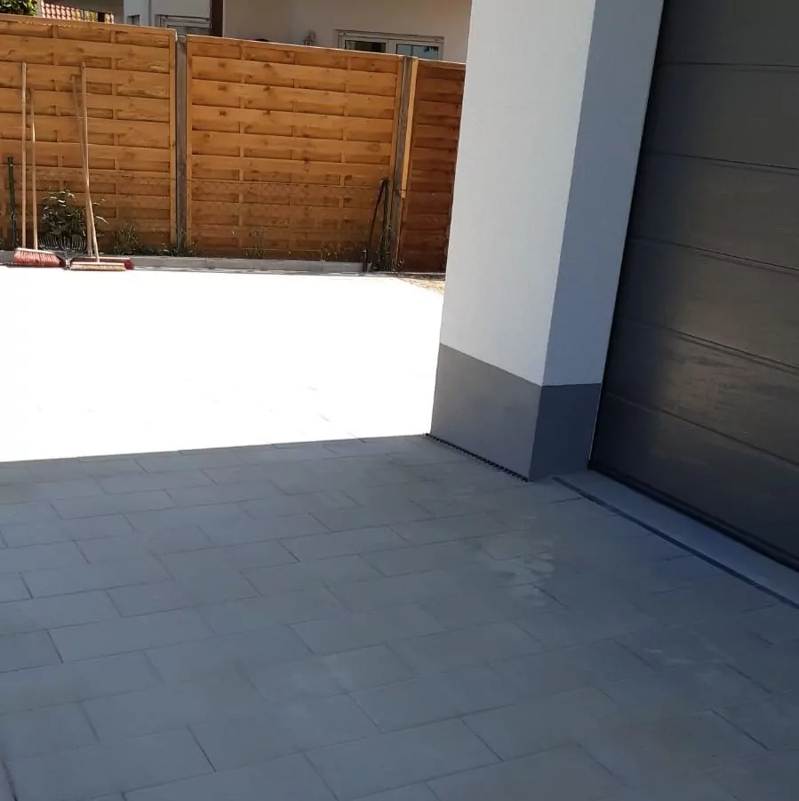 Eine mit hellgrauen Fliesen gepflasterte Auffahrt grenzt an eine Garage mit einer dunklen Tür. Im Hintergrund ist ein Holzzaun zu sehen, an dem ein Besen und andere Gartengeräte lehnen. Der Bereich ist gut vom Sonnenlicht beleuchtet.