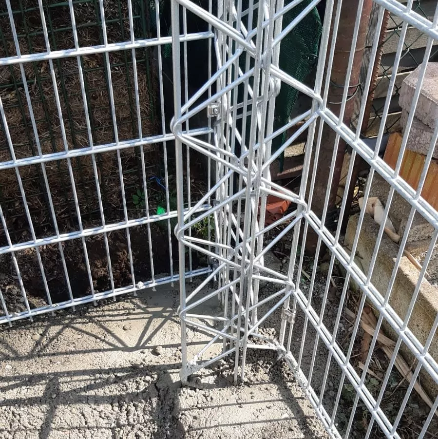 Eine Metallzaunecke aus verzinktem Stahlgitter ist an der Basis mit Beton befestigt. Die Struktur des Zauns umfasst vertikale und diagonale Stäbe zur Verstärkung, und in den umliegenden Bereichen sind Erde und Bauschutt zu sehen.