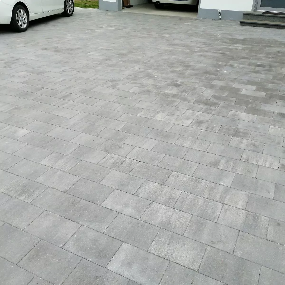 Eine Auffahrt besteht aus gleichmäßig verlegten grauen Pflastersteinen. Zwei weiße Autos sind zu sehen; eines ist teilweise zu sehen und ein anderes steht in einer Garage. Der Bereich ist sauber und gut gepflegt.