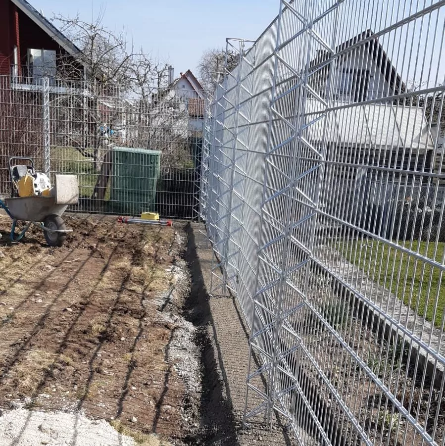 Entlang einer Baustelle ist ein Metallzaun mit Maschendrahtpaneelen und Geländer aufgestellt. Links ist eine Schubkarre zu sehen, im Vordergrund eine Mischung aus Gras und Erde. Im Hintergrund sind Häuser und ein blattloser Baum zu sehen.