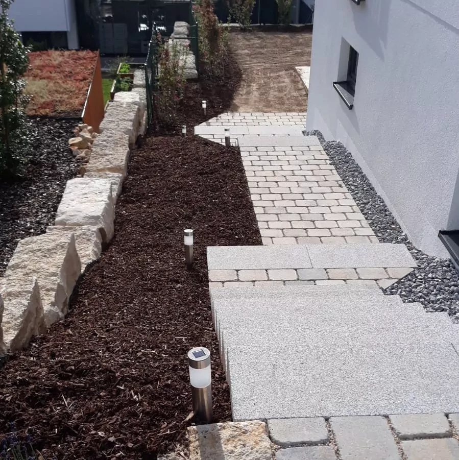 Ein mit Steinfliesen ausgelegter und von niedrigen Steinmauern gesäumter Gartenweg. Auf einer Seite befinden sich Mulch und kleine Sträucher, während solarbetriebene Lampen gleichmäßig entlang des Weges verteilt sind. Auf der rechten Seite ist ein Gebäude mit weißen Wänden zu sehen.