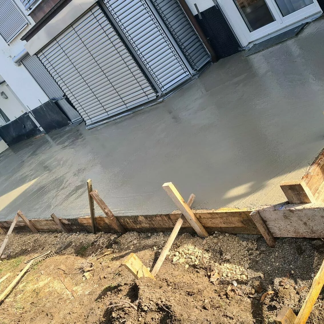 Frisch gegossene Betonplatte vor einem Gebäude mit geschlossenen Fensterläden. Holzbretter sind als temporärer Rahmen um den Umfang herum angebracht. Der Bereich scheint Teil laufender Bau- oder Renovierungsarbeiten zu sein.