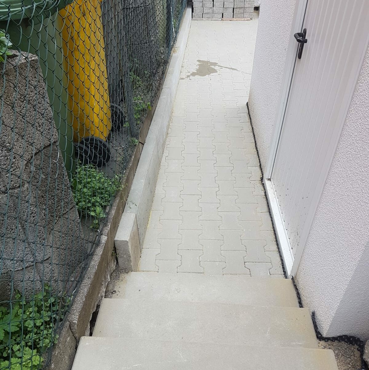 Weg aus Verbundpflaster zu einer weißen Tür, links ein grüner Zaun