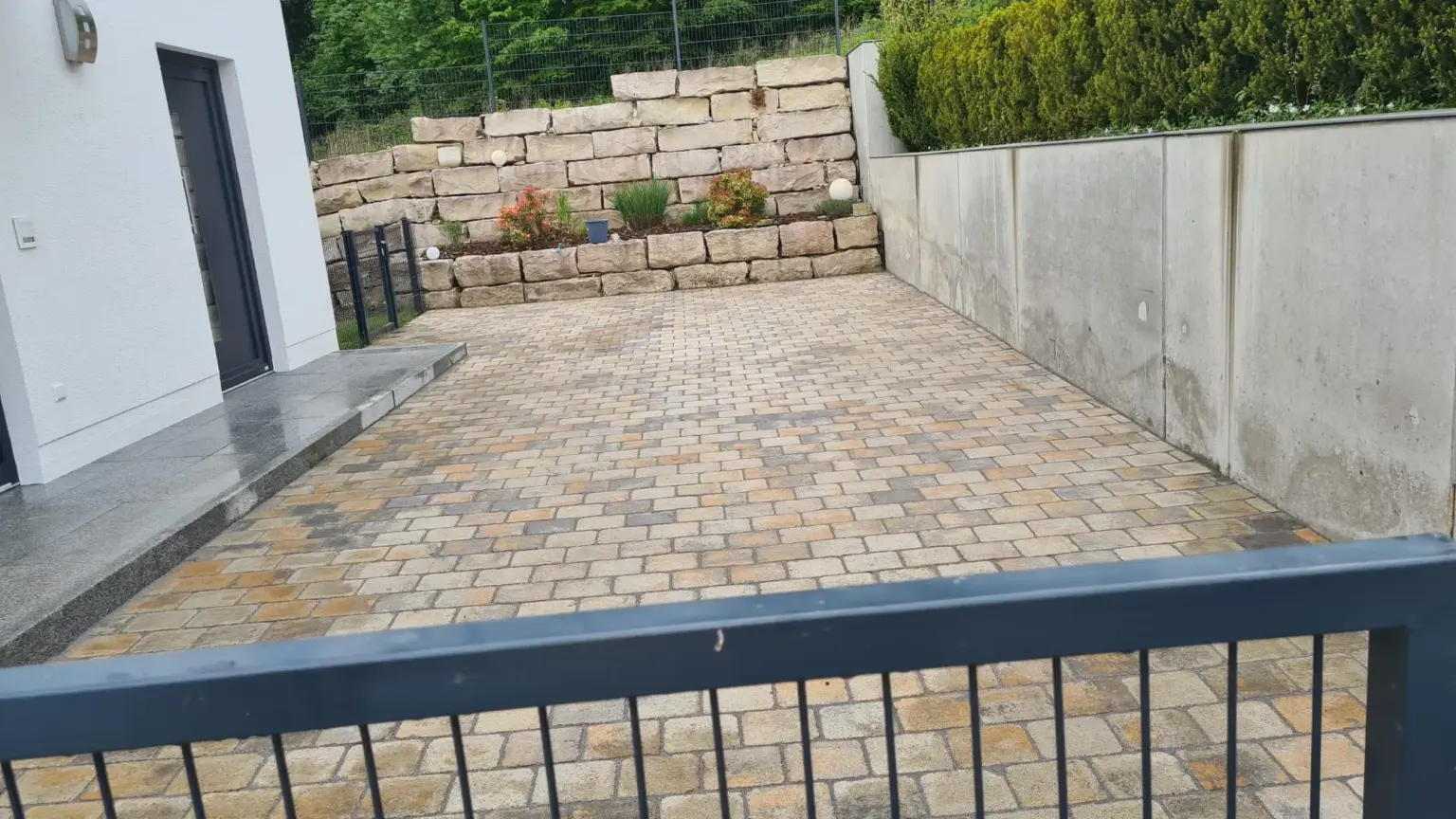 Eine mit Verbundsteinen gepflasterte Auffahrt führt zu einem geschlossenen schwarzen Metalltor. Links ist die Ecke eines weißen Gebäudes und rechts eine begrünte Betonmauer. Am Ende sind bepflanzte Steinstützmauern zu sehen.
