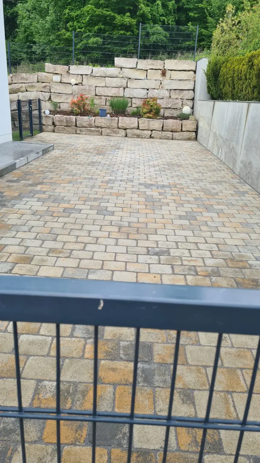 Eine mit Verbundsteinen gepflasterte Auffahrt führt zu einer Betonmauer. Hinter der Mauer befindet sich ein Gartenbeet mit verschiedenen Pflanzen und einer Stützmauer aus Stein. Im Vordergrund ist ein Metallzaun zu sehen, im Hintergrund sind Bäume zu sehen.