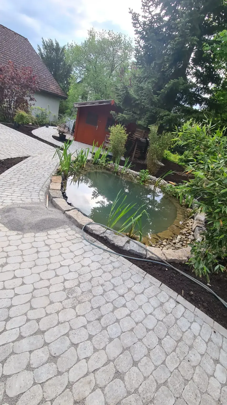 In der Nähe eines roten Holzschuppens befindet sich ein kleiner Teich, der von Steinen und Pflanzen umgeben ist. Der Bereich wird von einem Kopfsteinpflasterweg begrenzt, Bäume und Büsche bilden eine üppige Kulisse.