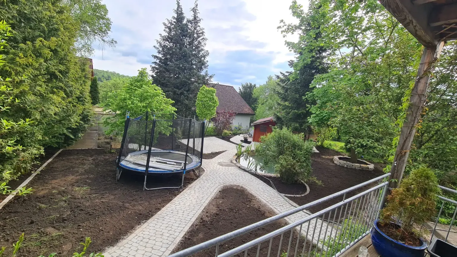 Ein Garten im Hinterhof mit einem Trampolin mit Sicherheitsnetz auf einem gepflasterten Steinweg. Der Hof ist von grünen Bäumen und Sträuchern umgeben. Im Hintergrund ist unter einem bewölkten Himmel ein kleines Haus zu sehen.