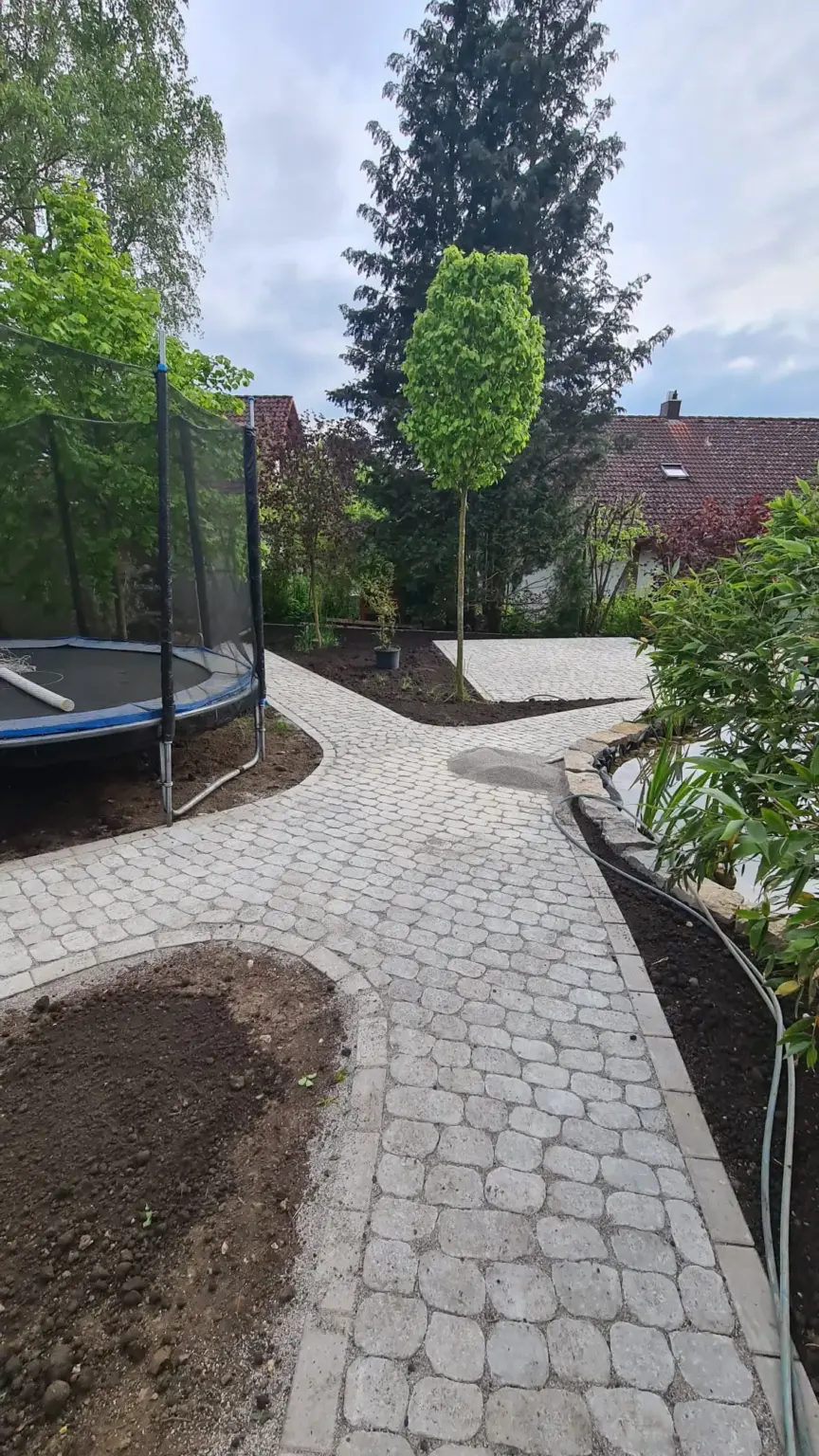 Ein Hinterhof mit einem gepflasterten Steinweg, der sich um ein Trampolin und frisch bepflanzte Gartenbeete windet. Rechts steht ein junger Baum, und im Hintergrund ist neben einem kleinen Haus ein großer immergrüner Baum zu sehen. Am Himmel ist der Himmel bedeckt.