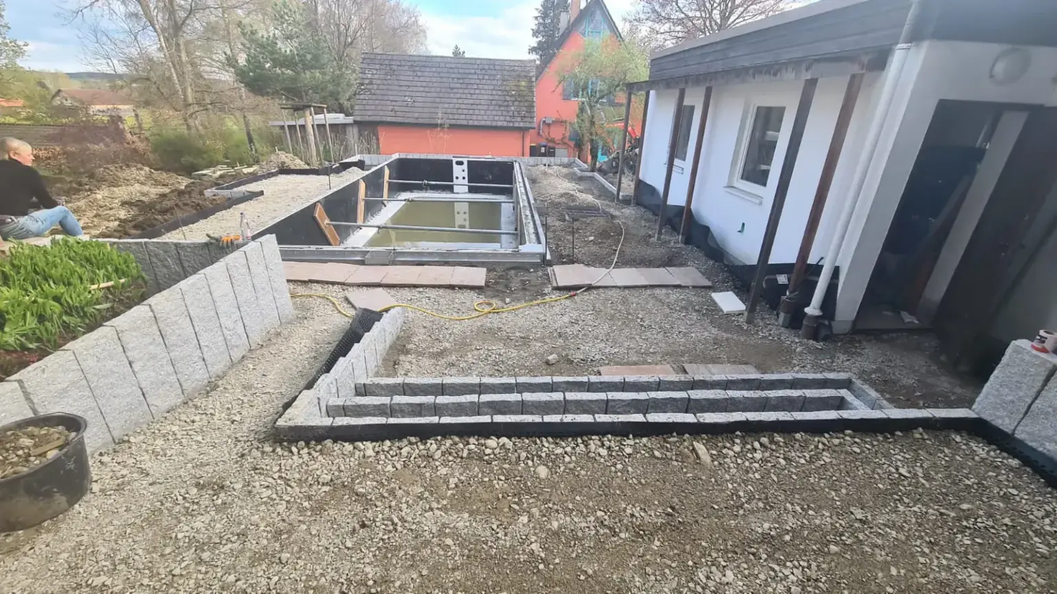 Eine Baustelle mit einem teilweise fertiggestellten Swimmingpool, umgeben von Kies und Betonblöcken. In der Nähe steht ein kleines weißes Gebäude mit dunklem Dach, links sitzt eine Person auf dem Boden. Im Hintergrund sind Bäume und ein rotes Gebäude zu sehen.