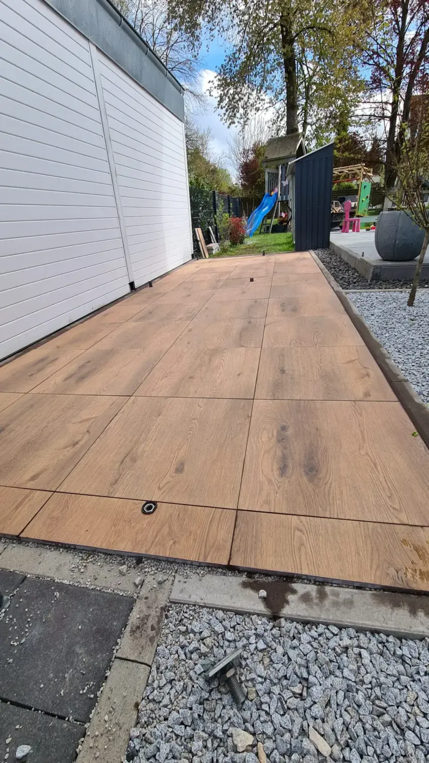 Neben einer weißen Garage ist ein neu installiertes Holzdeck zu sehen. Das Deck ist von Kies umgeben und im Hintergrund ist ein teilweise sichtbarer Garten mit Bäumen und Spielgeräten zu sehen. Auf der Oberfläche des Decks ist eine kleine schwarze Halterung zu sehen.