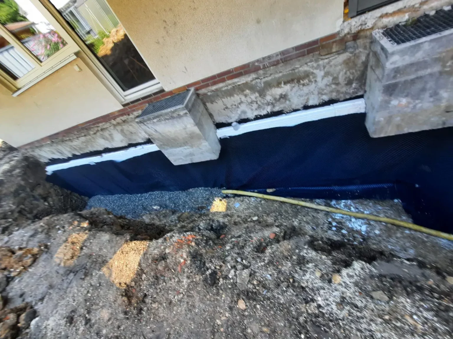 Ein Hausfundament ist freigelegt, mit Schichten aus Kies und einer wasserdichten Membran. Zwei Betonblöcke sind an der Wand befestigt. In der Umgebung ist ausgehobene Erde zu sehen, und auf dem Boden ist ein gelber Schlauch zu sehen.