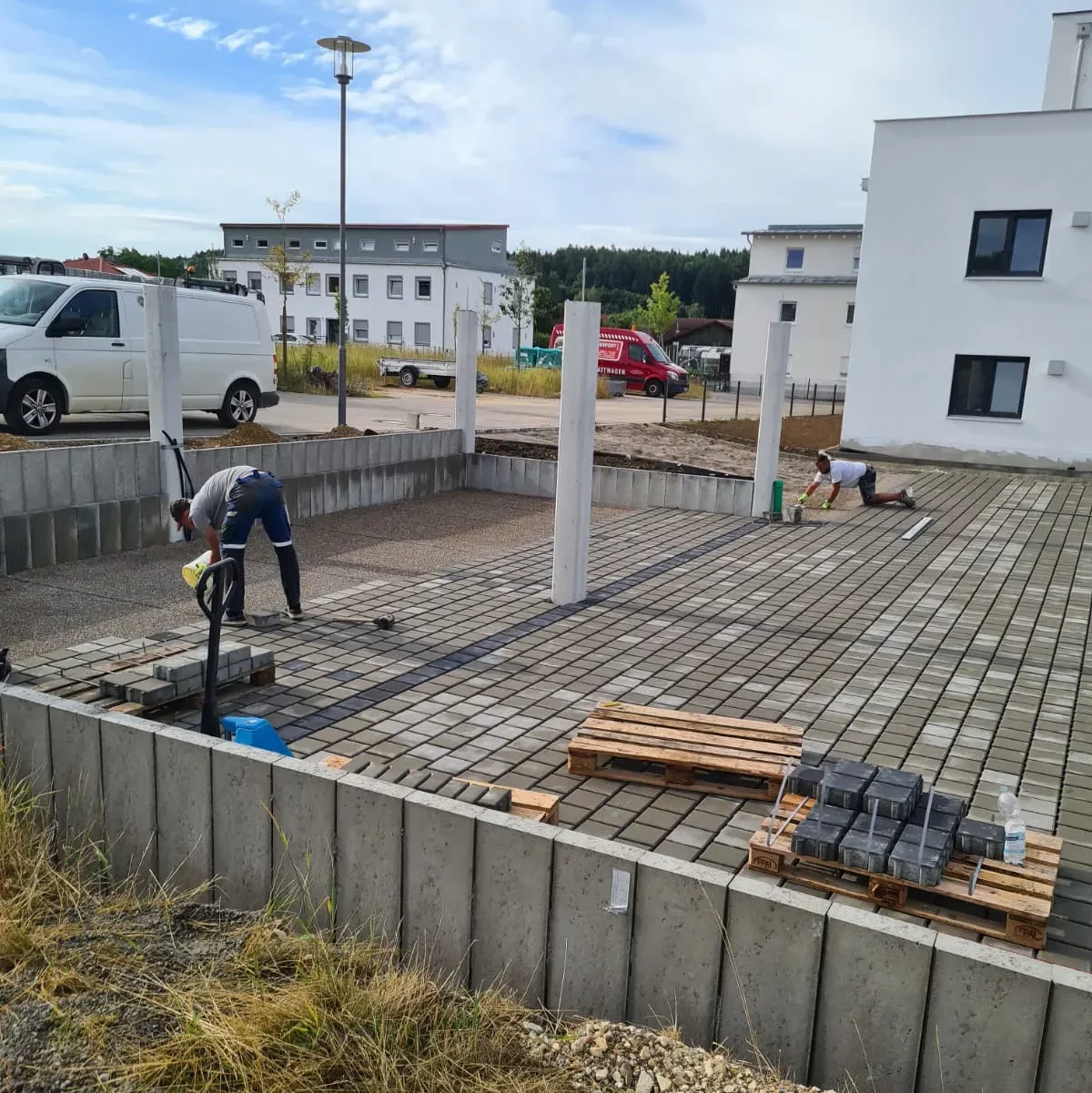 Zwei Arbeiter verlegen Steinfliesen in einem von Betonmauern begrenzten Außenbereich. In der Nähe parken ein weißer Lieferwagen und ein rotes Auto. Im Hintergrund ist ein modernes weißes Gebäude zu sehen, im Vordergrund eine Rasenfläche.